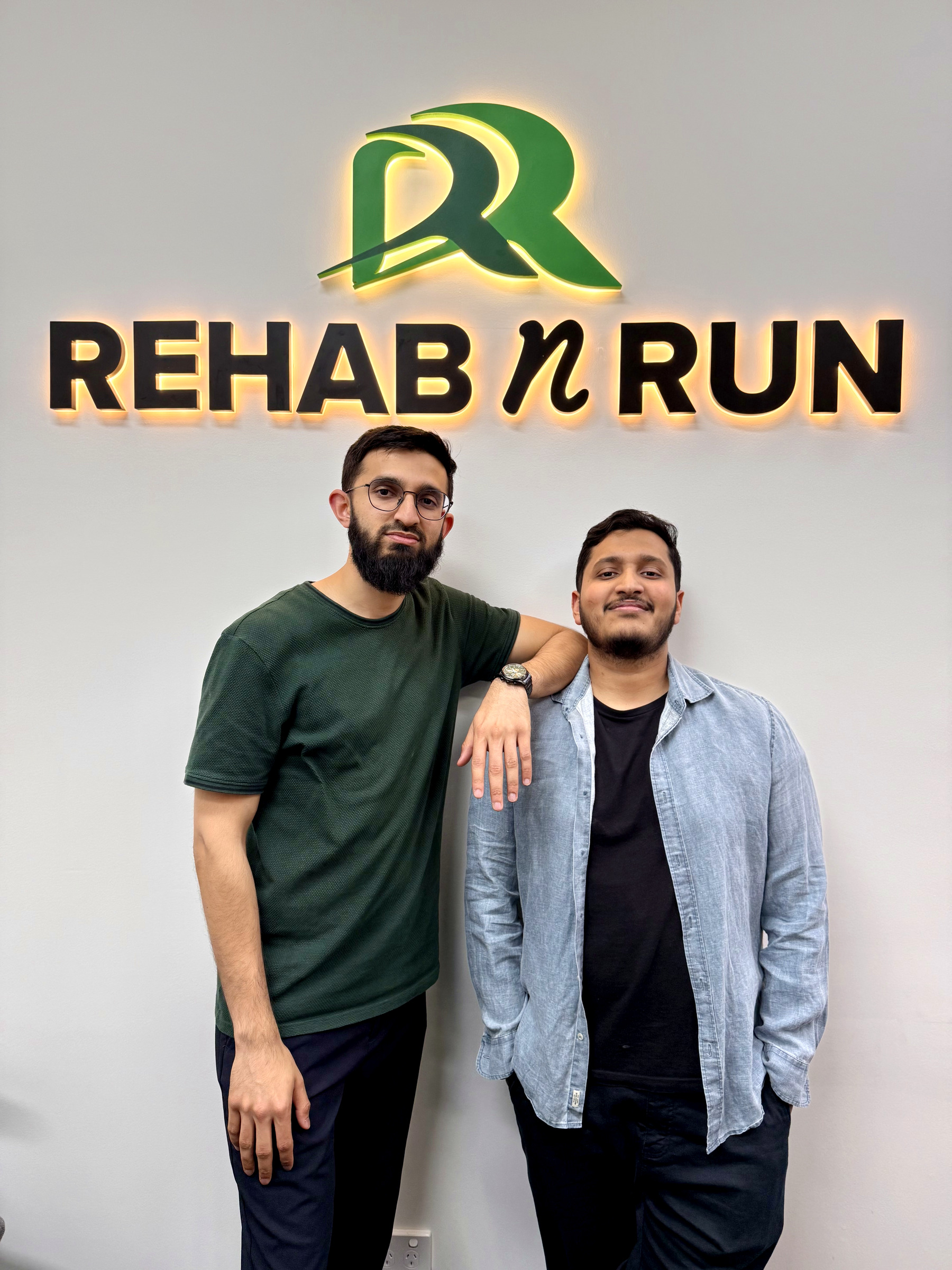 Abdulla Omar and Fawad Faheem standing beneath the illuminated Rehab n Run logo inside the Liverpool clinic